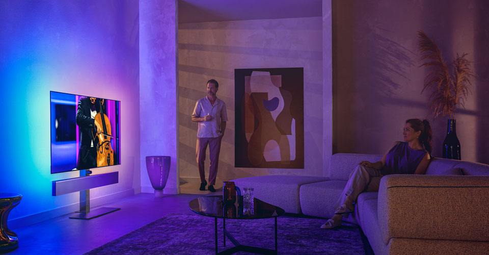 Vyras ir moteris stilingoje svetainėje žiūri &bdquo;Philips Ambilight&ldquo; televizorių, kuriame rodomas violončelės pasirodymas