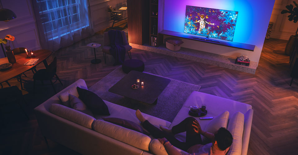 Vyras atsipalaiduoja ant sofos pritemdytame svetainėje ir žiūri &bdquo;Philips Ambilight&ldquo; televizorių