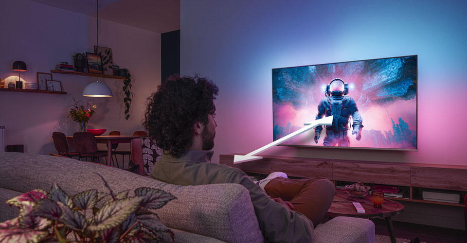 Vyras sėdi ant sofos ir žiūri &bdquo;Philips Ambilight&ldquo; televizorių, kuriame rodomas spalvingas astronauto vaizdas su rodykle, rodančia optimalų žiūrėjimo atstumą